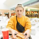 Przystojny młody mężczyzna ubrany w czapkę i żółte ubrania siedzi w recepcji i jedzenie fast food