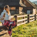 Rano dziewczyna siedzi na drewnianej ławce w górach na łonie natury, czyta książkę i pije gorącą herbatę z termosu. Pojęcia Czytanie w naturze. Trening na zewnątrz