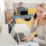 Całkiem uśmiechnięta młoda bizneswoman pracuje na laptopie i rozmawia przez telefon przy biurku w sypialni, jej mąż siedzi z laptopem na łóżku