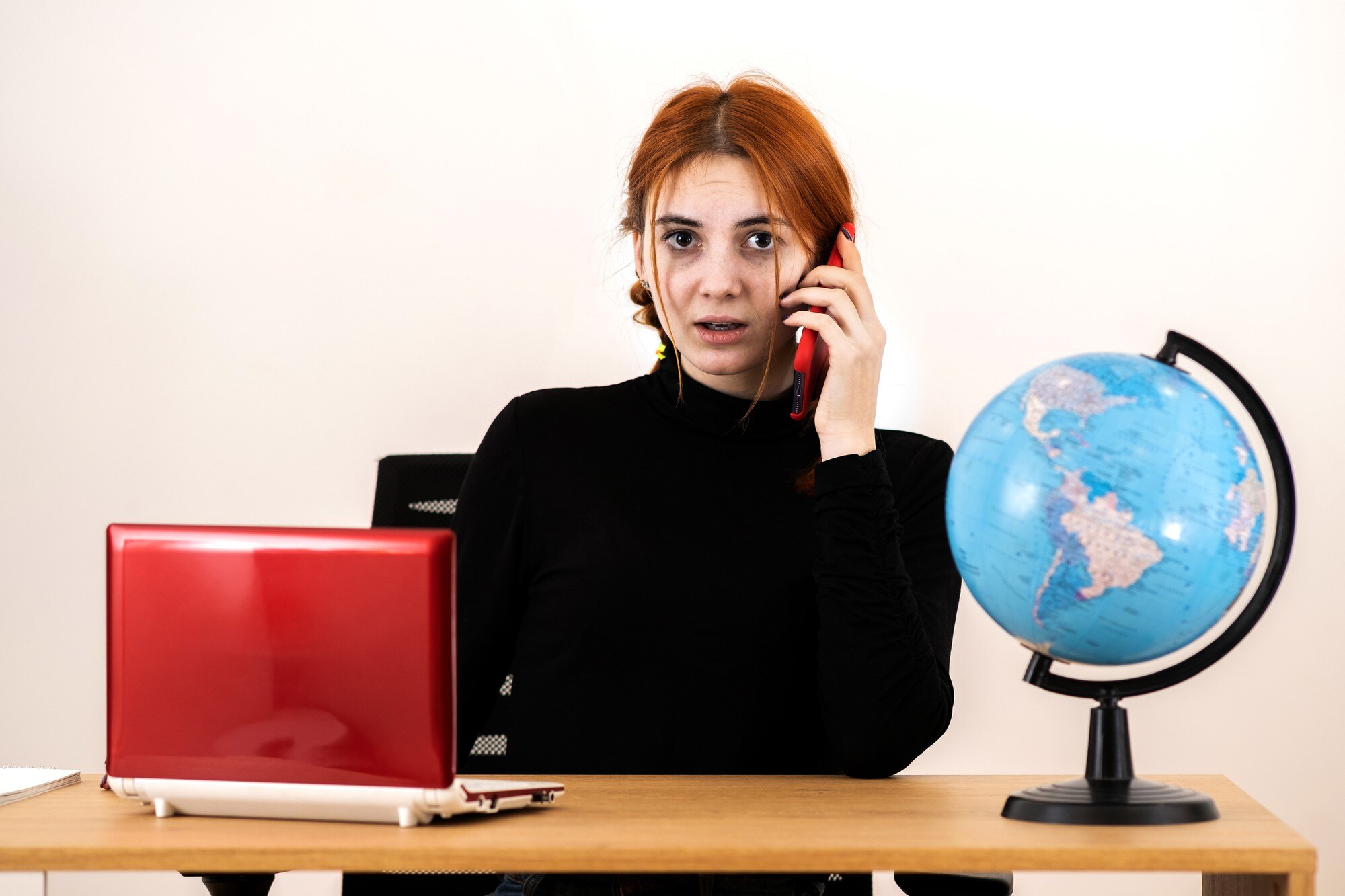 Młoda kobieta biura podróży siedzi za biurkiem z laptopa i globu geograficznego świata rozmawia przez telefon komórkowy.