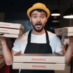 Sprzedawca pizzy trzyma kartonowe pudełka z przekąskami, wygląda z ekspresją omg, nosi żółty kapelusz i biały t