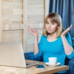 Niepewny! Portret zdezorientowanej młodej dziewczyny freelancer z blond fryzurą bob w niebieskiej koszulce siedzi w kawiarni i pracuje na laptopie z uniesionymi rękami i zdziwioną twarzą, patrząc w kamerę. wnętrz