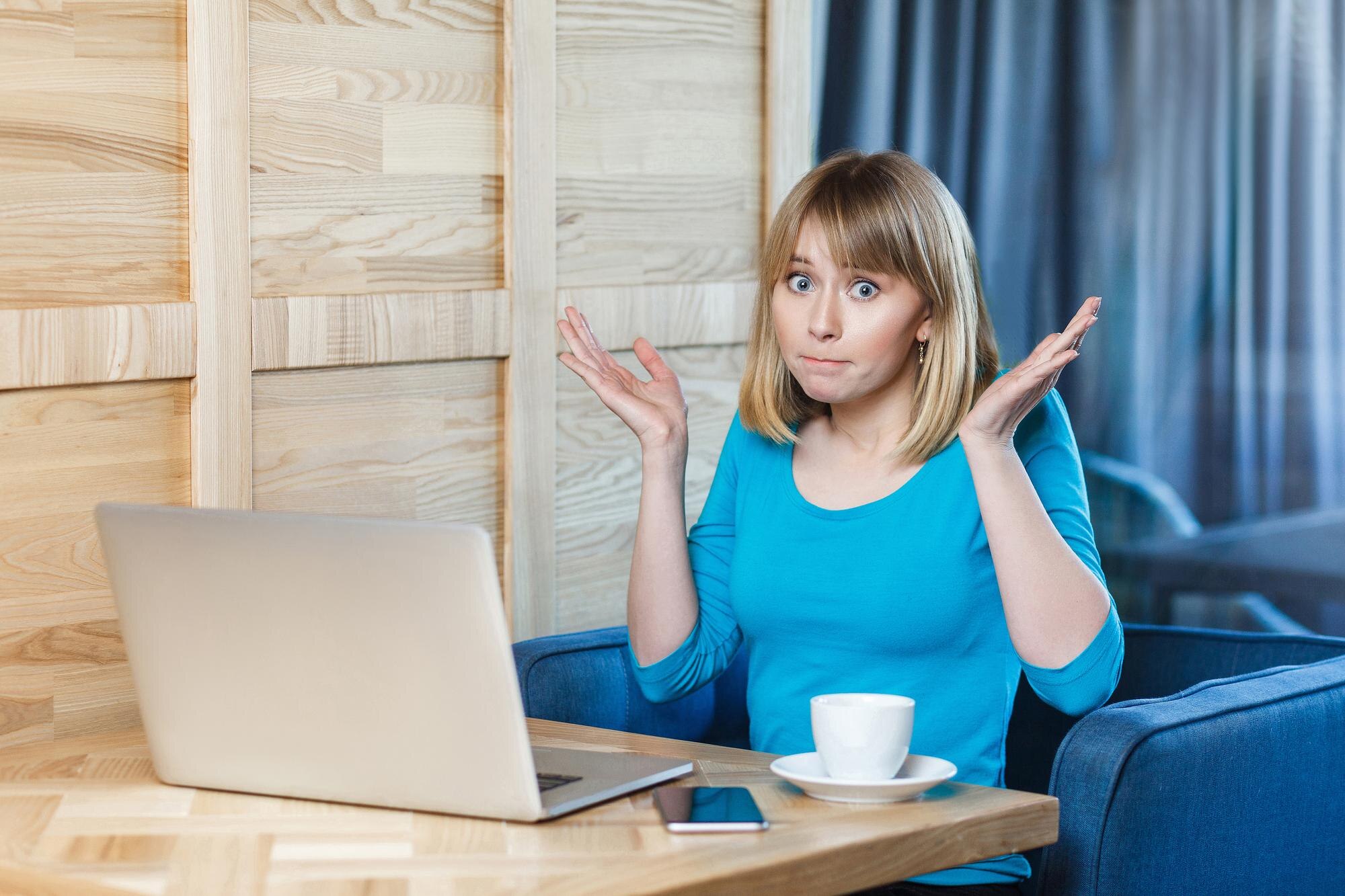 Niepewny! Portret zdezorientowanej młodej dziewczyny freelancer z blond fryzurą bob w niebieskiej koszulce siedzi w kawiarni i pracuje na laptopie z uniesionymi rękami i zdziwioną twarzą, patrząc w kamerę. wnętrz