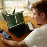 Osoba przy drewnianym biurku zajmuje się intensywnym wielozadaniowością z dwoma smartfonami wyświetlającymi wykresy analizę danych kodowanie na laptopie scena nowoczesnej wydajności w środowisku pracy technologicznej