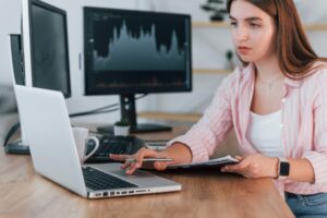 Korzystając z laptopa Kobieta makler giełdowy w zwykłych ubraniach pracuje w biurze za pomocą komputera