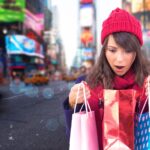 Zszokowana brunetka otwierająca torbę z prezentami przed rozmytą ulicą Nowego Jorku