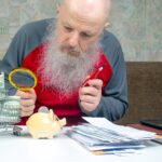 starszy mężczyzna w czerwonej koszuli używając szkła powiększającego do dokładnego zbadania jego pieniędzy pokazujących finansowe