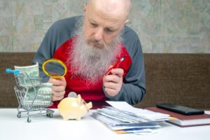 starszy mężczyzna w czerwonej koszuli używając szkła powiększającego do dokładnego zbadania jego pieniędzy pokazujących finansowe