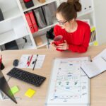 Młoda dziewczyna siedzi przy biurku w biurze, trzymając w dłoniach czerwony kubek i patrząc na monitor. Tablica magnetyczna leży przed dziewczyną.