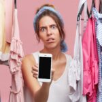 Wściekła kobieta o zmęczonym wyglądzie, stojąca między ubraniami, pokazująca telefon komórkowy stojąc w szatni. Młoda kobieta z wyrazem niezadowolenia robi zakupy, używając inteligentnego telefonu w pomieszczeniu
