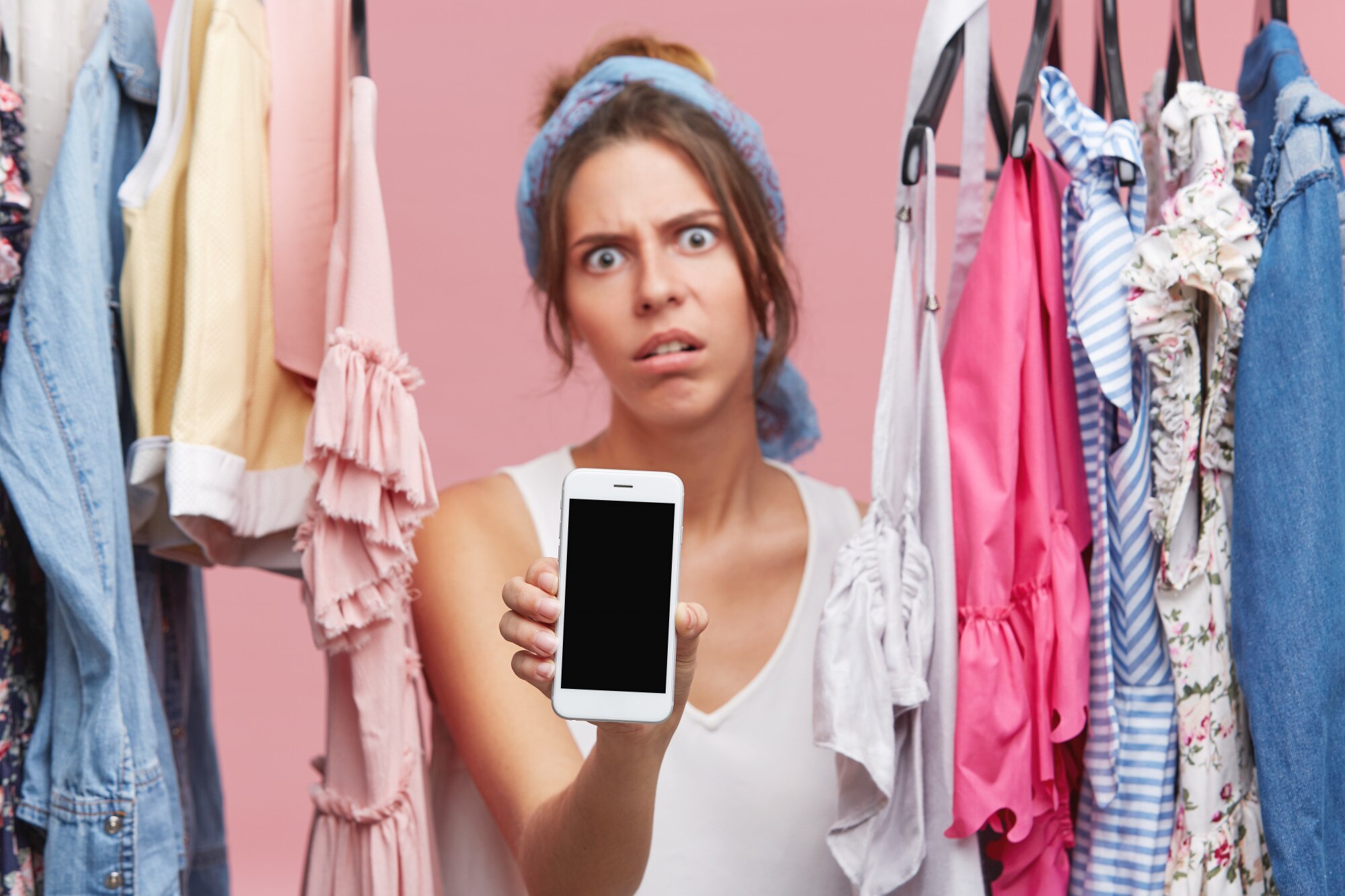 Wściekła kobieta o zmęczonym wyglądzie, stojąca między ubraniami, pokazująca telefon komórkowy stojąc w szatni. Młoda kobieta z wyrazem niezadowolenia robi zakupy, używając inteligentnego telefonu w pomieszczeniu